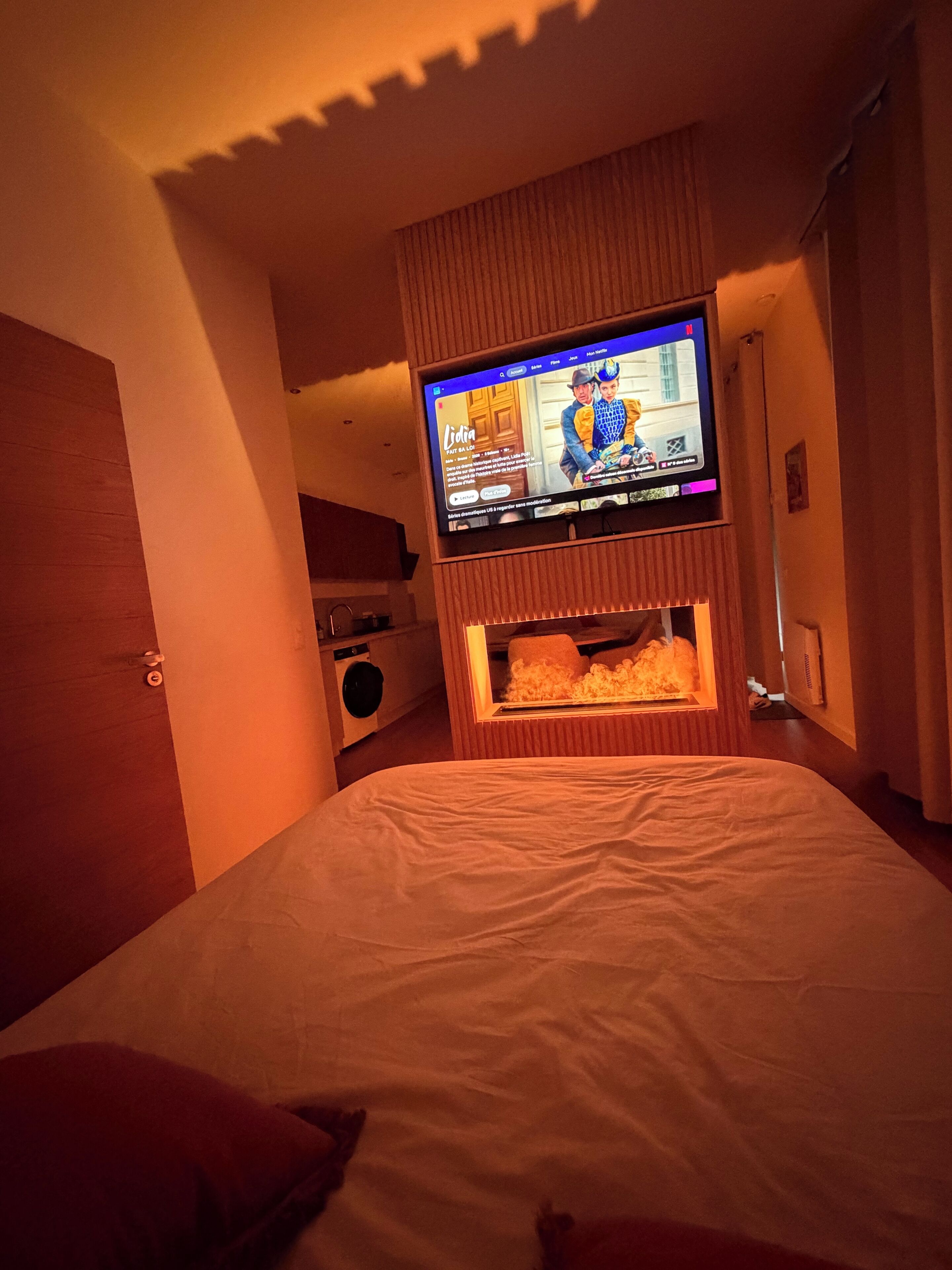 A hotel room with a bed, a television mounted on a wooden panel, and a washing machine in the background.