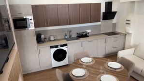 Uma cozinha compacta com micro-ondas, cafeteira e máquina de lavar roupa. Possui uma área de refeições com mesa posta para quatro pessoas.