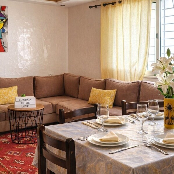 Un comedor con una mesa dispuesta para cuatro personas, un sofá, una mesita auxiliar y un jarrón decorativo con flores.