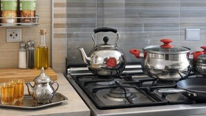 Una cucina con piano cottura a gas, bollitore, pentola e un vassoio con tazze e teiera. Si notano contenitori per spezie e un cesto di vimini a parete.