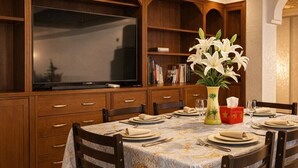 Zona pranzo con un tavolo rotondo apparecchiato per sei persone, mobili in legno, televisore e un vaso con fiori bianchi.