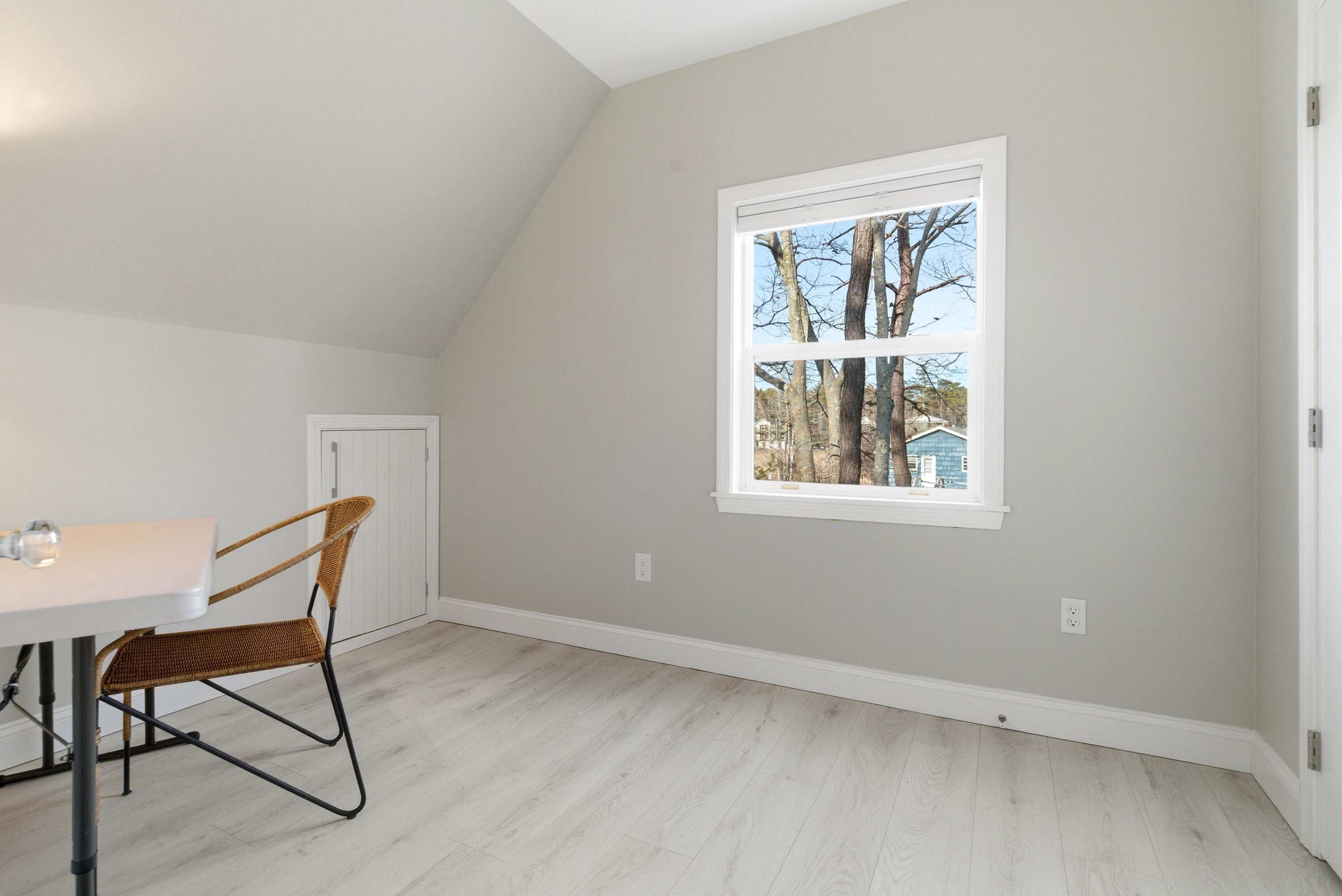 A room with a sloped ceiling, a window with a view of trees and a house, a white door, and a dining table with a chair.