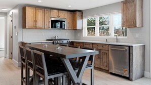 A modern kitchen with wooden cabinets, a stainless steel dishwasher, and a large island with chairs. - Drift Cottage Steps to Beach (Old Orchard Beach)