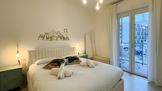 Ein Hotelzimmer mit Bett, Nachttisch, Wandlampe, Fenster mit Blick auf Gebäude sowie einer Wanddekoration von Bergen und Bäumen.
