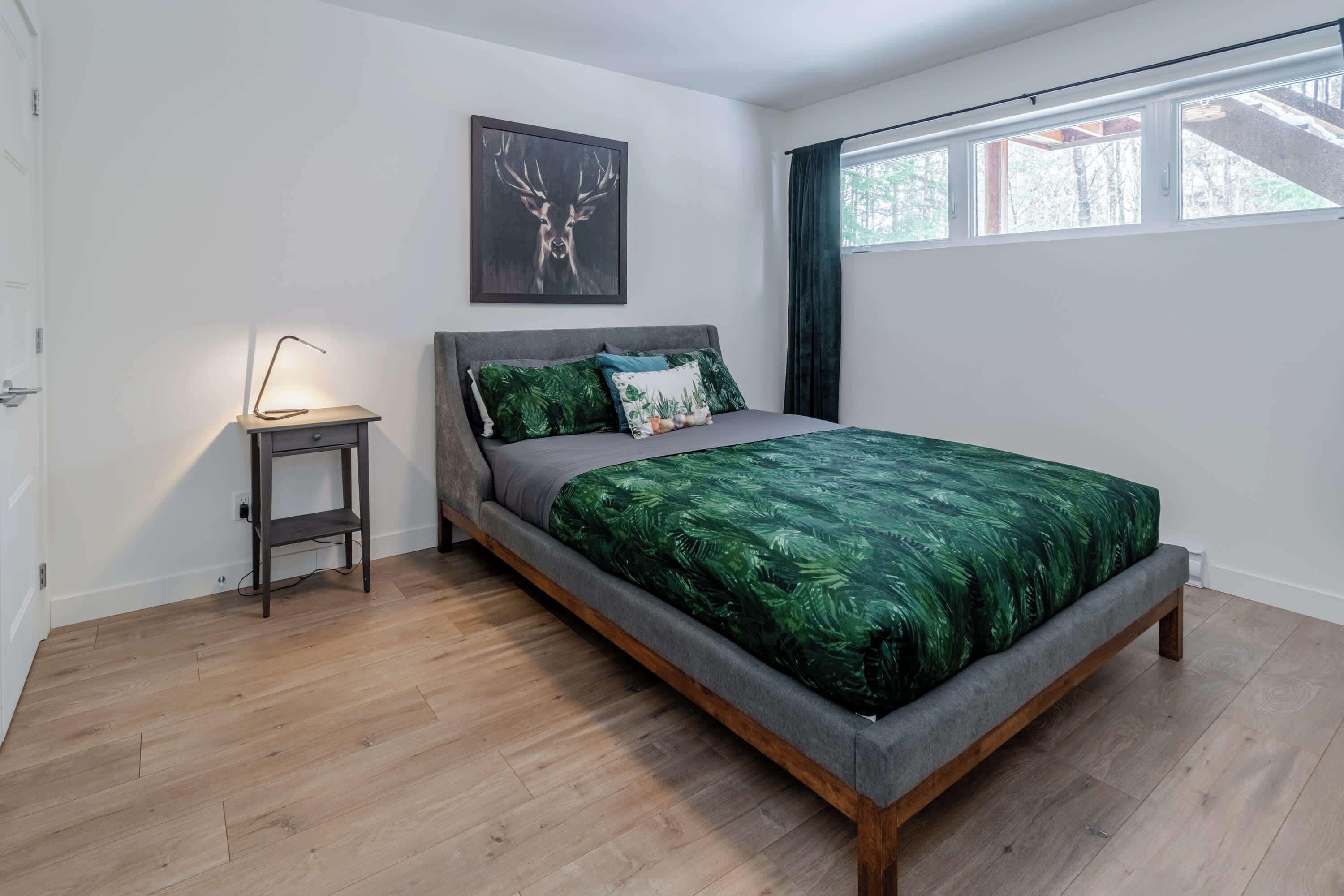 Ein Schlafzimmer mit Bett, Nachttisch, Lampe, Fenster und einem Wandbild.