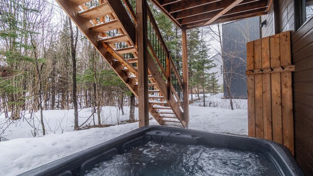 Una vasca idromassaggio con un pontile in legno e scale che conducono a un edificio, immersa in una foresta innevata.