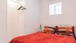 A bedroom with a red bedspread, a white wardrobe, a small window, and a wall-mounted light fixture. - Apartment for rent at Rua de Santo Estevão 25, Santa Maria Maior, Lisbon. (Lisboa)