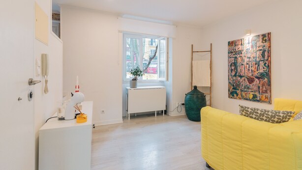 A modern living room with a yellow sofa, a radiator, a potted plant, and a large abstract painting on the wall. - Apartment for rent at Rua de Santo Estevão 25, Santa Maria Maior, Lisbon. (Lisboa)