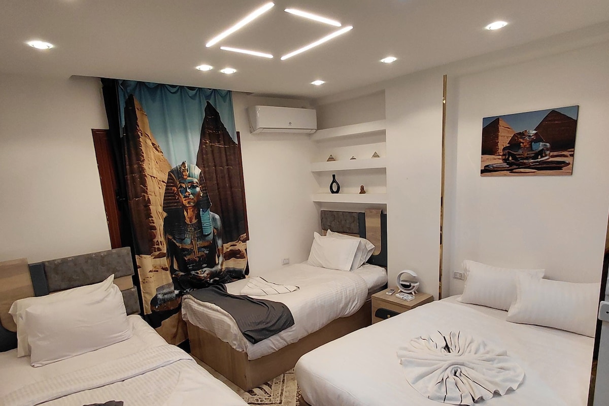 Quarto de hotel com duas camas, uma mesa de cabeceira, uma escrivaninha com chaleira e ar condicionado fixado na parede.