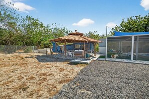 Un espacio de estar al aire libre cubierto con mesas y sillas, un jardín cercado y un edificio con fachada azul.
