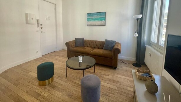 Ett vardagsrum med en brun soffa, ett runt soffbord, en turkosfärgad pall, en tavla på väggen och ett fönster med gardiner.