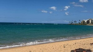 Une plage avec une eau d’un bleu limpide, un rivage sablonneux et un bâtiment à étages, avec des palmiers en arrière-plan.
