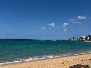 A beach with clear blue water, a sandy shore, and a building with multiple floors and palm trees in the background. - Lovely Beach Villa in Vega Alta Puerto Rico near to Dorado (Sabana, Vega Alta, Puerto Rico)