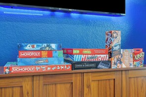 Verschiedene Brettspiele stehen auf einem Regal vor einer blauen Wand, darüber befindet sich ein Fernseher.