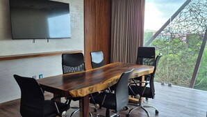 Un salón de conferencias con una gran mesa de madera, sillas de oficina negras, un televisor de pantalla plana montado en la pared y vistas a zonas verdes a través de un amplio ventanal.