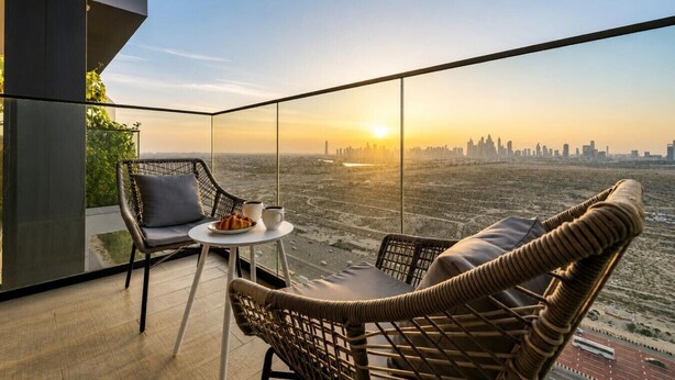 A balcony with wicker chairs, a small table, and a cityscape view at sunset.