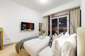 A hotel room with a bed, a TV mounted on the wall, a window with a city view, and a nightstand. - Luxe JVC Studio with Skyline Views, Pool and Gym (Dubai)