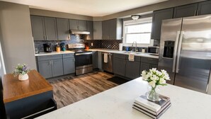 A modern kitchen with grey cabinets, stainless steel appliances, a marble countertop, and a wooden dining table. - Work + Relax + Play | 3BR Home w/ Ping Pong Table (Liverpool)
