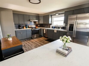 A modern kitchen with grey cabinets, stainless steel appliances, a marble countertop, and a wooden dining table. - Work + Relax + Play | 3BR Home w/ Ping Pong Table (Liverpool)