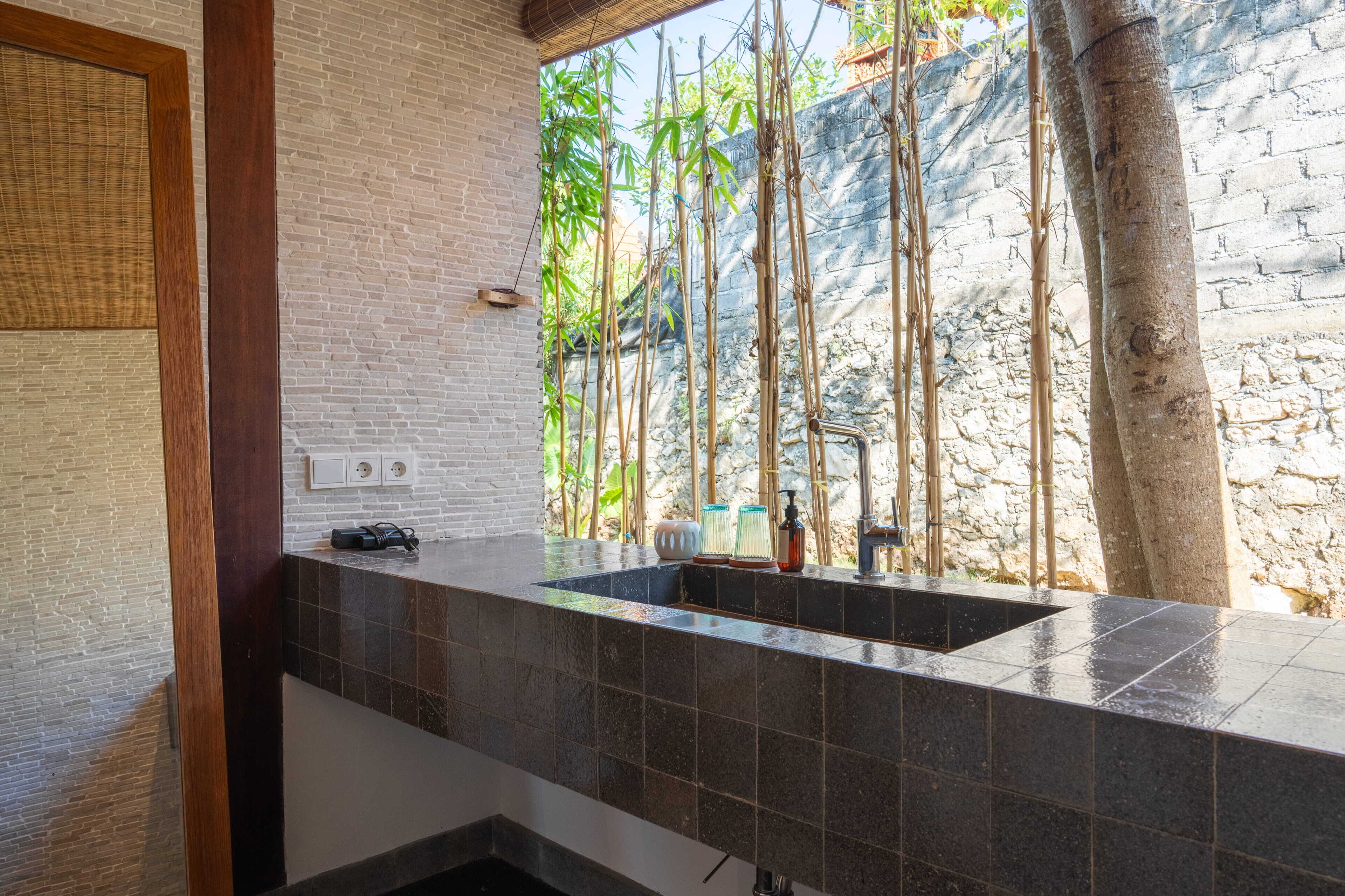 A bathroom with a large sink, a mirror, and a window overlooking bamboo and a stone wall.