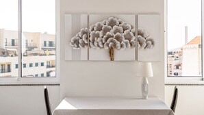 A dining area with a table, chairs, a wall-mounted artwork, and a window with a view of buildings. - Sé Cathedral Rooftop, a Home in Madeira (Funchal)