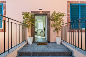 Eine Treppe führt zu einer Tür mit Fußmatte, gesäumt von Blumentöpfen und blauen Fensterläden.