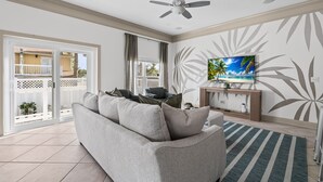 A living room with a grey sofa, a ceiling fan, a TV on a wooden stand, and a view through a sliding glass door to an outdoor area. - Villa De Le Mar - Pool, Movie Theater (Jacksonville Beach)