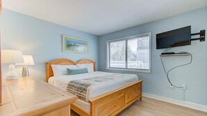 Een slaapkamer met een houten bed, een nachtkastje met een lamp, een raam met zonwering en een tv aan de muur.