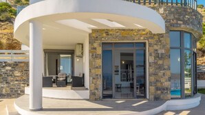 Eine moderne Villa mit geschwungenem Äußeren, einer Steinfassade und einer Glastür, die zu einem Innenbereich mit Blick auf einen Pool führt.