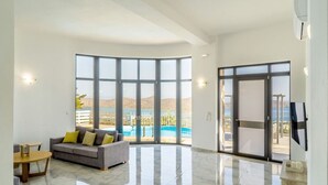 Ein großzügiger Wohnbereich mit einem großen Fenster, einem grauen Sofa mit gelben Kissen, einem Holztisch und einem Pool, der durch das Fenster sichtbar ist.