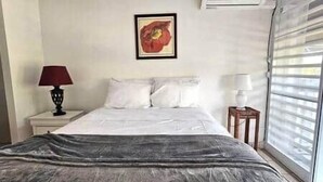 Ein Schlafzimmer mit einem Bett, zwei Nachttischen, einem Fenster mit Jalousien und einer Wand-Klimaanlage.