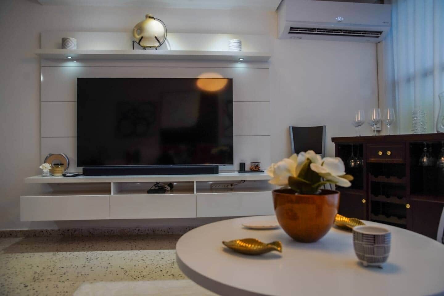 Una sala moderna con un televisor de pantalla plana grande montado en una pared blanca, una mesa de centro redonda con objetos decorativos y un mueble de madera oscura con copas de vino.