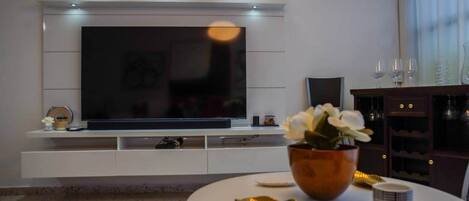 Una sala moderna con un televisor de pantalla plana grande montado en una pared blanca, una mesa de centro redonda con objetos decorativos y un mueble de madera oscura con copas de vino.