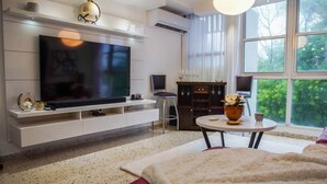 A modern living room with a large flat-screen TV, a fireplace, a dining area, and a large window with a view of trees. - Prime Location in Condado Puerto Rico (San Juan)