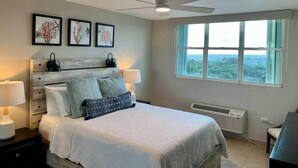 A bedroom with a bed, bedside lamps, a ceiling fan, a window with a view, and framed artwork on the wall. - P1207-Oceanview Apartment- Peñamar (Fajardo)