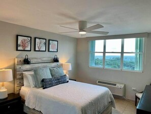 Ein Schlafzimmer mit Bett, Nachttischlampen, Deckenventilator, Fenster mit Blick und gerahmten Kunstwerken an der Wand.