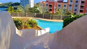 A swimming pool with a curved edge, surrounded by greenery and a fence. - P1207-Oceanview Apartment- Peñamar (Fajardo)