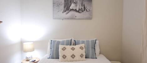 A bedroom with a bed, pillows, a nightstand, a lamp, and a wall-mounted picture.