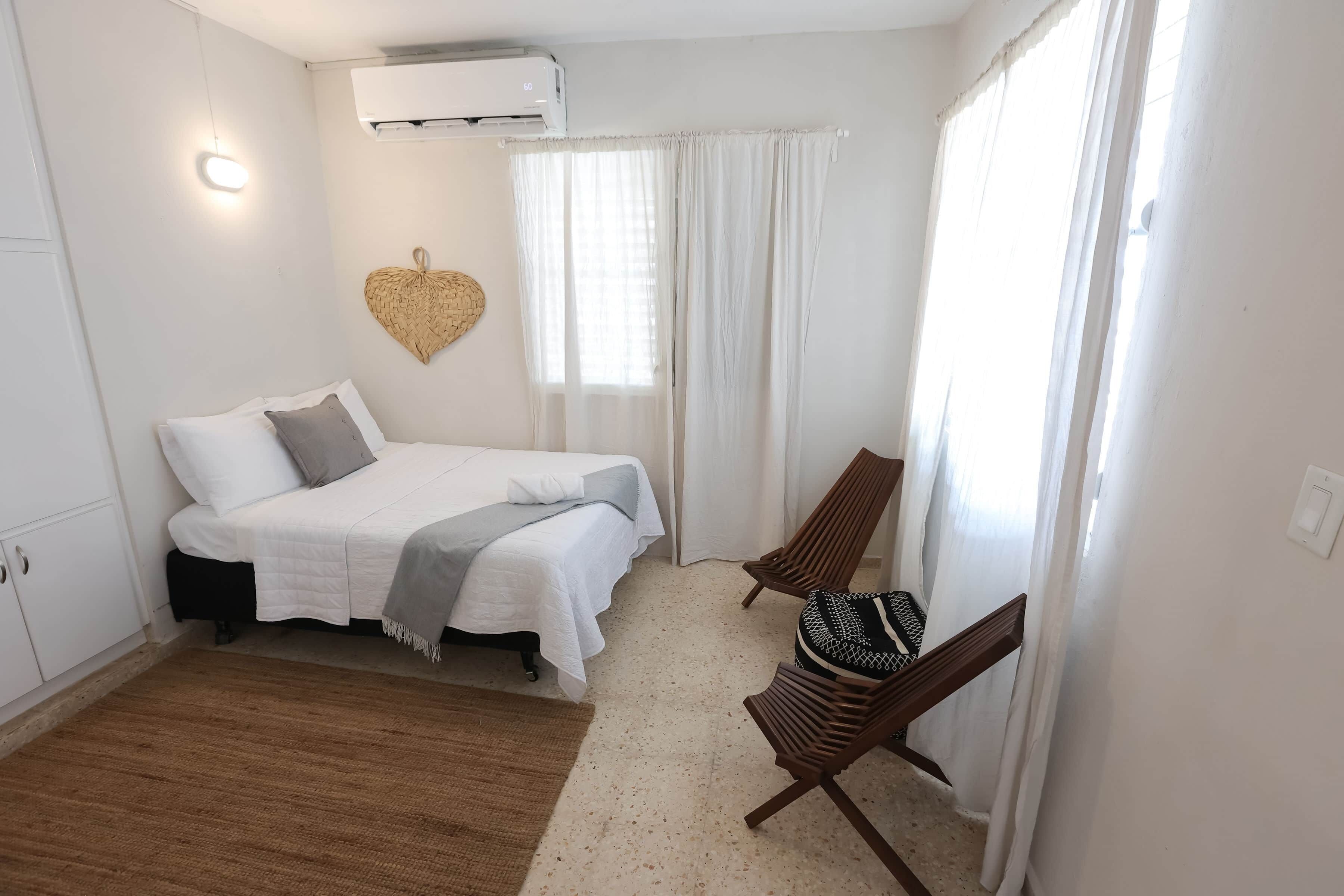 A bedroom with a bed, a chair, a window with curtains, an air conditioner, and a wall-mounted light.