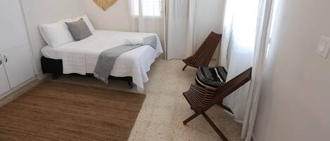 A bedroom with a bed, a chair, a window with curtains, an air conditioner, and a wall-mounted light.