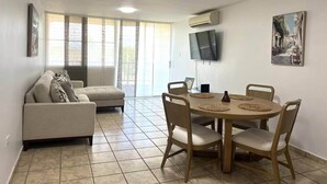 Un salon comprenant un canapé, une table à manger avec des chaises, un téléviseur à écran plat et une grande fenêtre avec des rideaux.