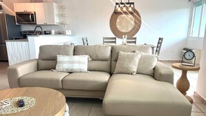 Un soggiorno con un divano angolare beige, un tavolino rotondo, una zona pranzo con tavolo e sedie, una cucina con elettrodomestici moderni e un'ampia decorazione circolare a parete.