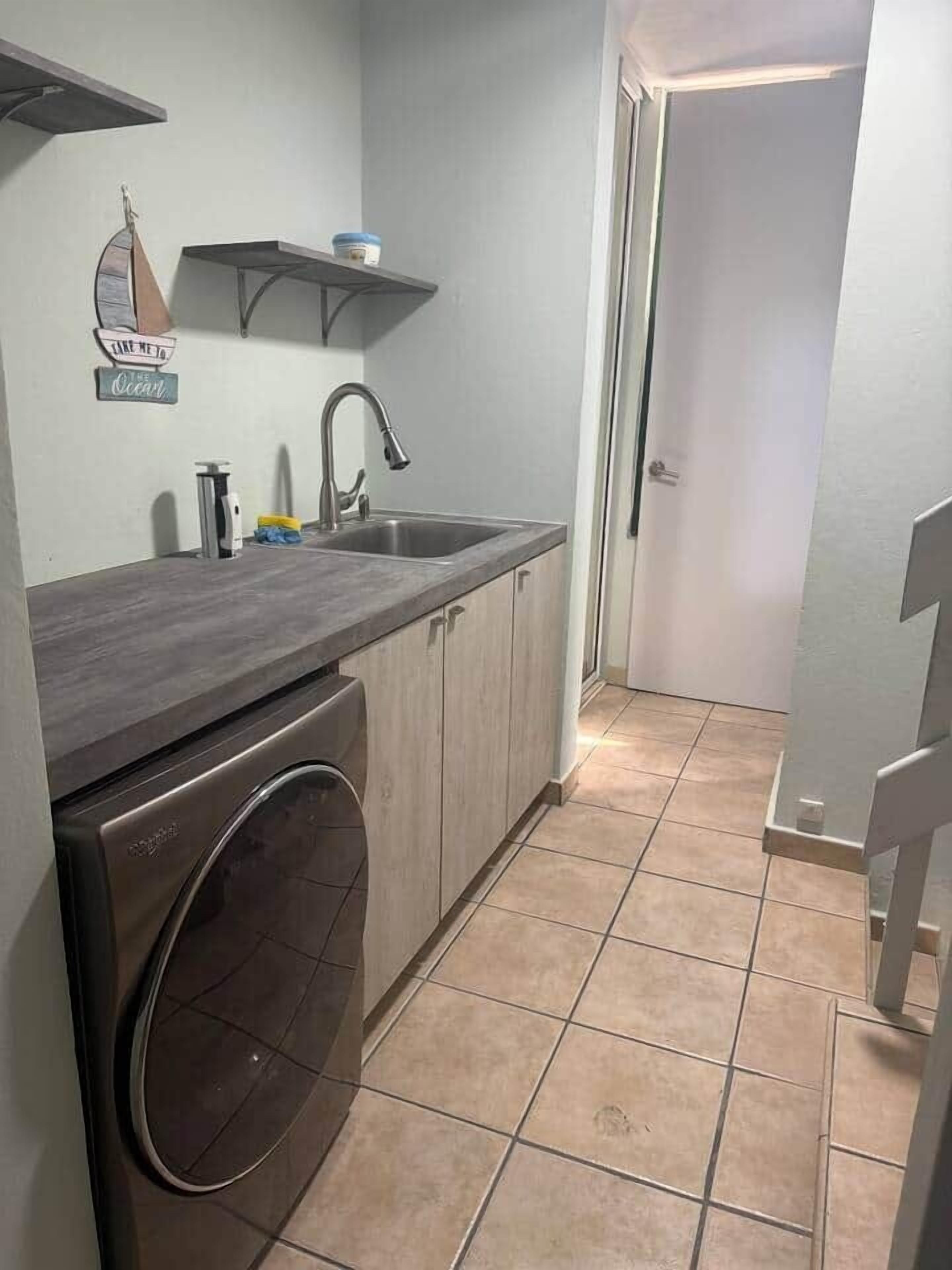 Ett litet kök med tvättmaskin, diskho och en dörr som leder till ett annat rum.