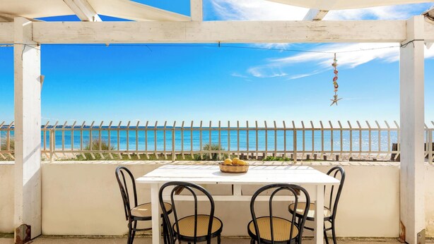 Ein überdachter Außenbereich mit weißem Tisch und Stühlen, mit Blick auf einen Strand, eine Umzäunung und einen klaren Himmel.