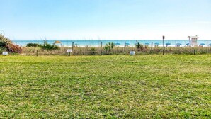 Un champ d’herbe avec une plage en arrière-plan, une tour de surveillance des sauveteurs et des parasols de plage.