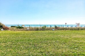 Un champ d’herbe avec une plage en arrière-plan, une tour de surveillance des sauveteurs et des parasols de plage.