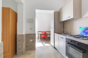 A compact kitchen with a gas stove, oven, and a dining area with a red chair. - Nella Suites - Bilo PT (Porto Cesareo)