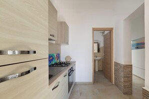 A modern kitchen with wooden cabinets, a gas stove, and a view into the adjacent bathroom. - Nella Suites - Bilo PT (Porto Cesareo)