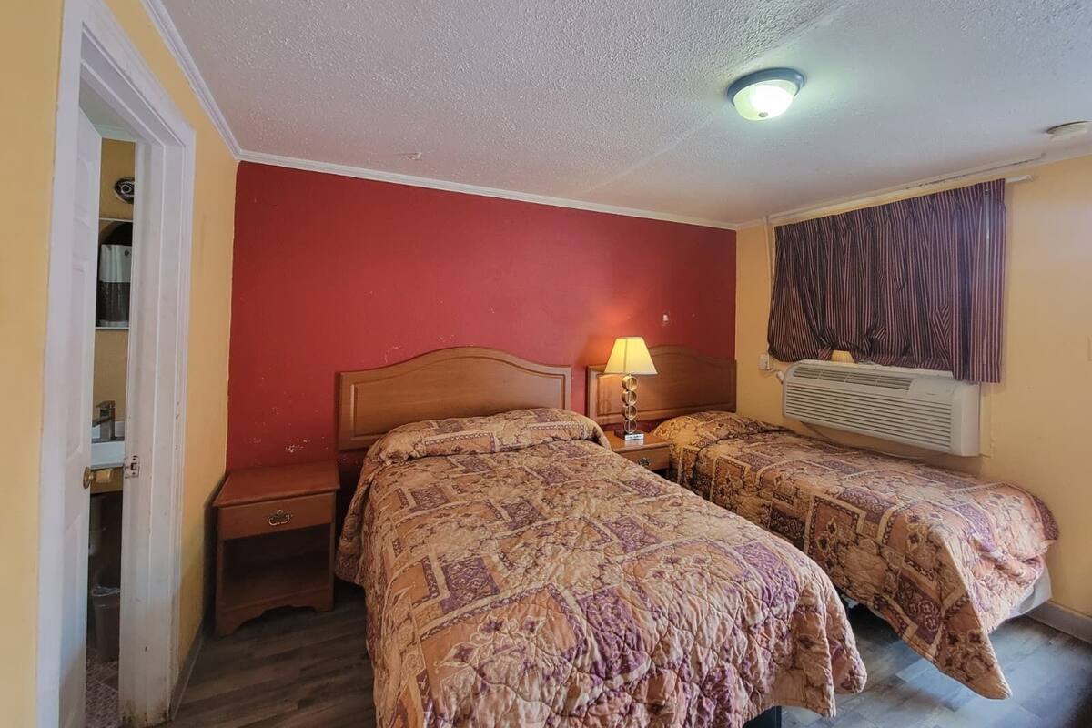 Habitación de hotel con dos camas, una mesita de noche, una lámpara, un equipo de aire acondicionado y una ventana con cortinas.
