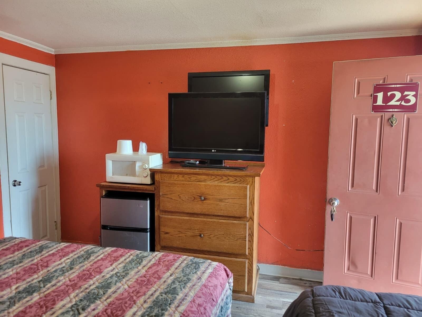 A bedroom with a bed, a dresser, a television, a microwave, and a refrigerator.